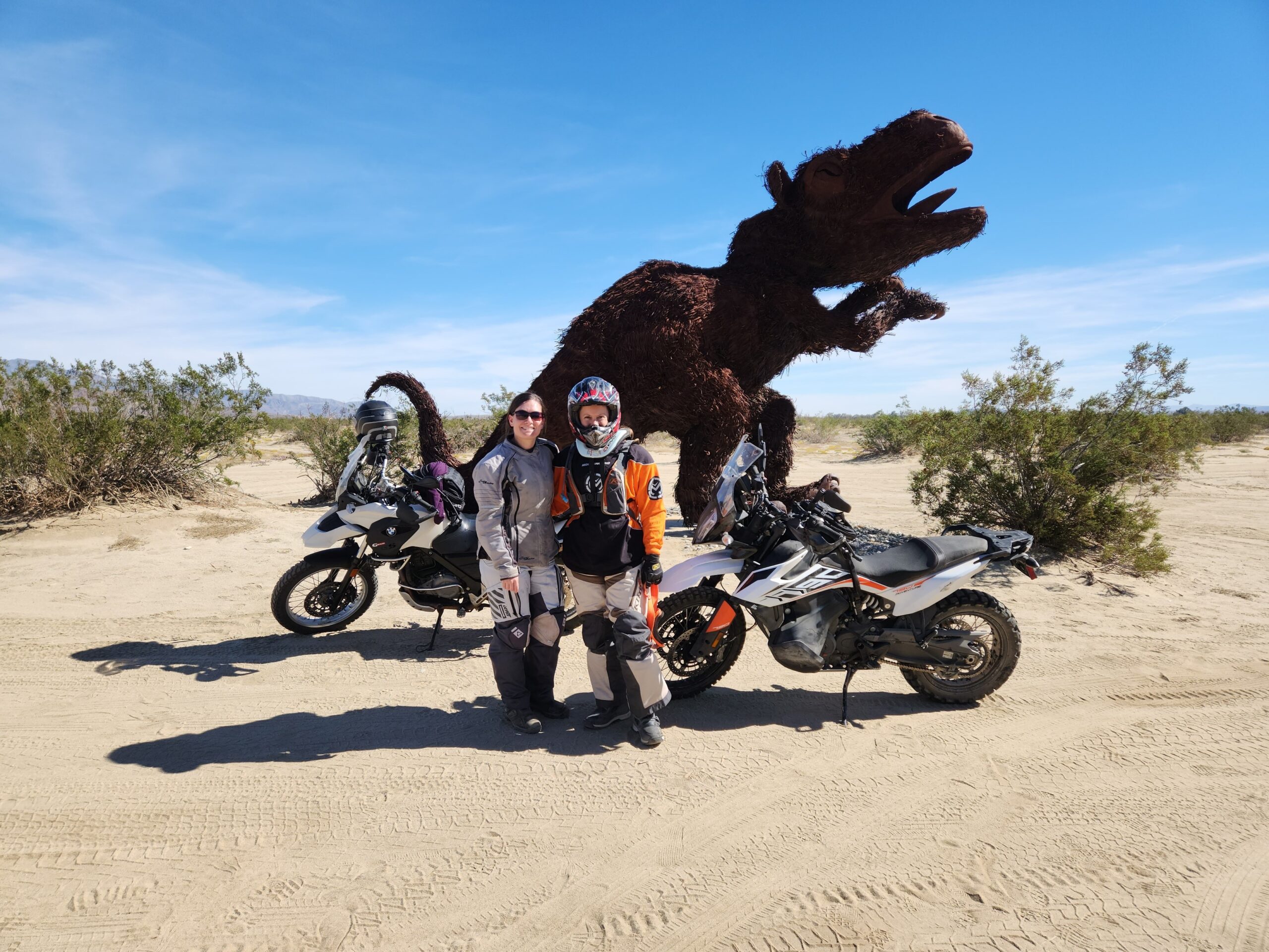 Two motorcycle riders, a KTM 790 adventure motorcycle, and a BMW G650GS adventure motorcycle, stand in front of a T-rex iron sculpture in Anza Borrego State Park, California