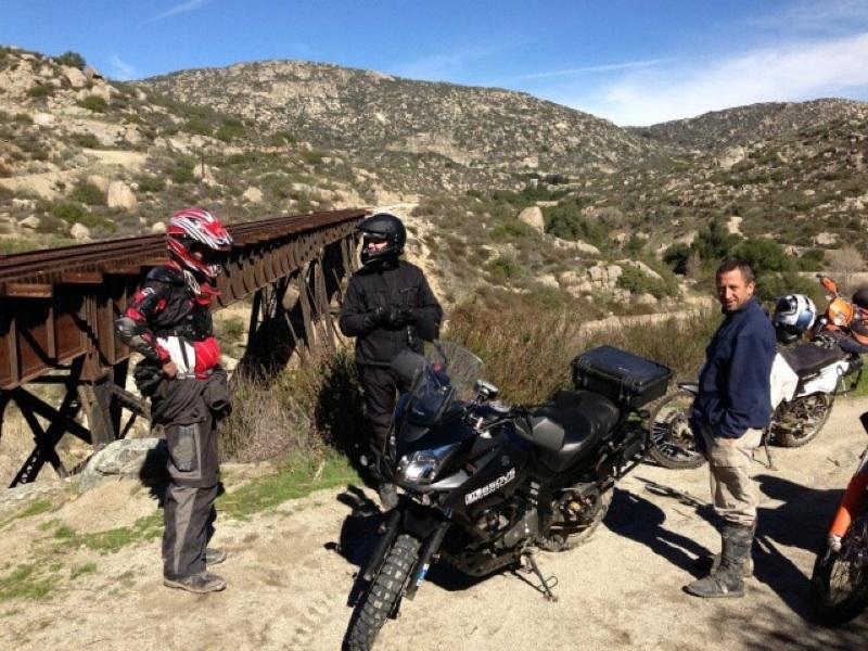 Rider near Suzuki V-strom adventure motorcycle talks to other motorcycle rider in front of old train trestle bridge outside of San Diego, California