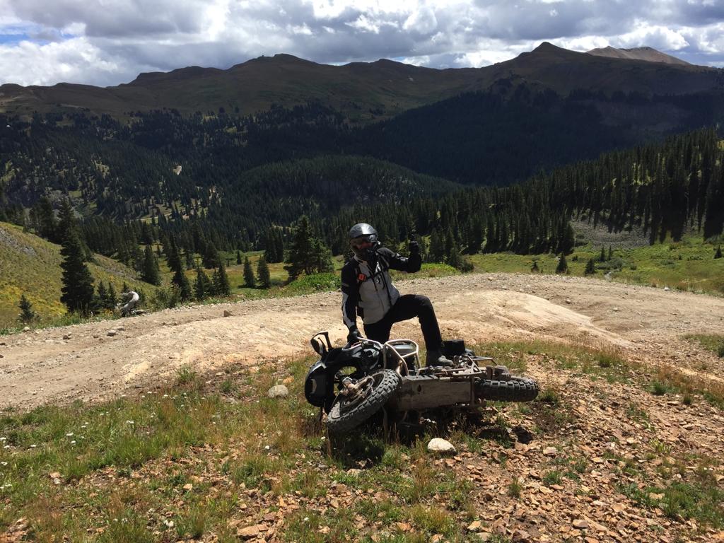 Motorcycle rider does Captain Morgan pose on top of fallen BMW 1200 GS adventure motorcycle on a dirt trail in the San Juan mountains of Southwestern Colorado