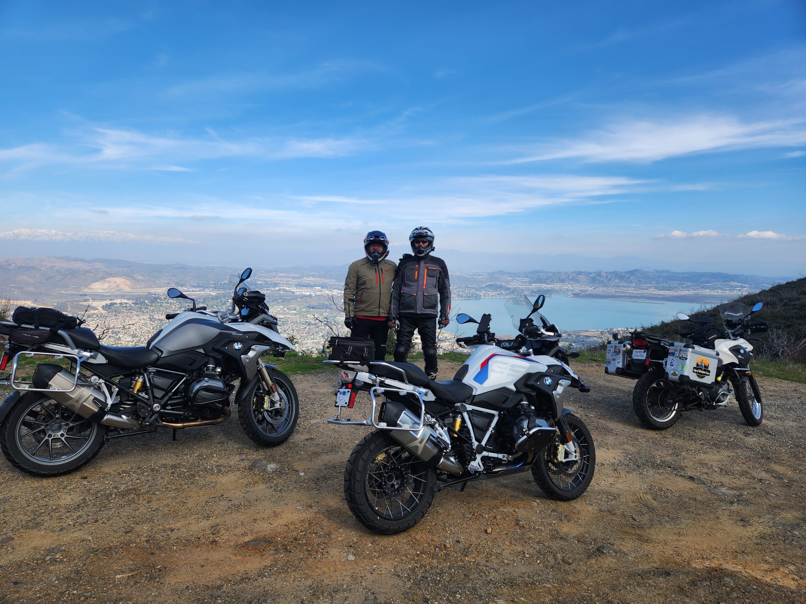 Two motorcycle riders stand on top of a mountain with 3 motorcycles in the foreground, with a lake in the background