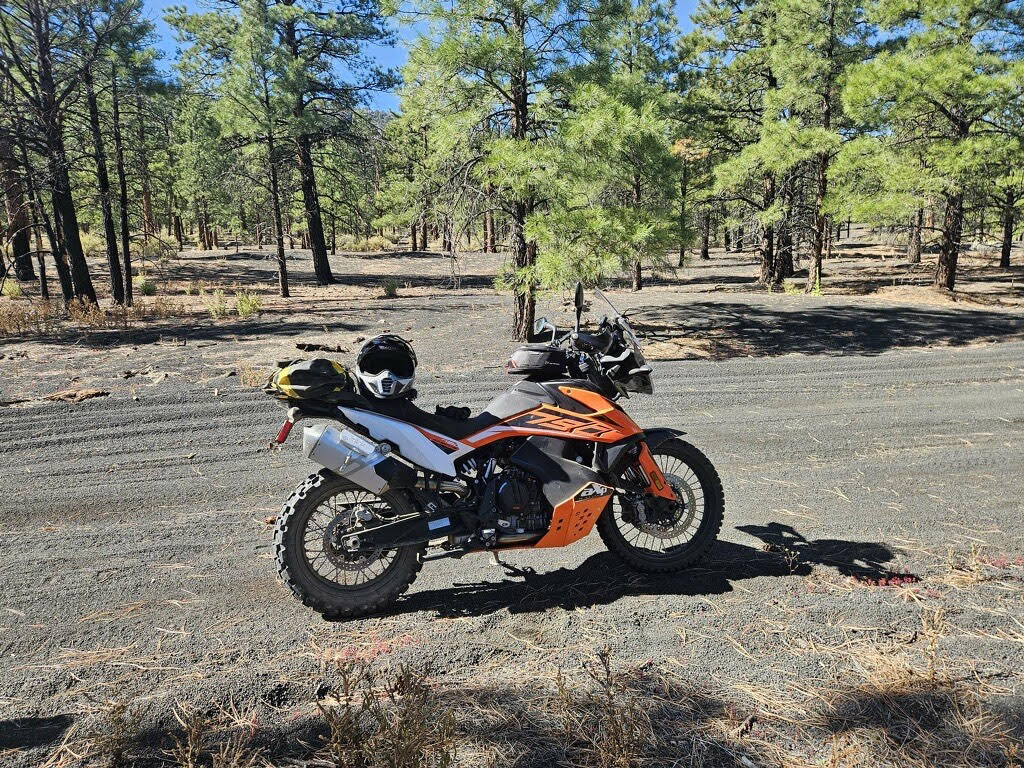 KTM 790 Adventure motorcycle is pictured on a trail in Cinders Off-Highway Vehicle Area with an ash road surface and pine forest in the background