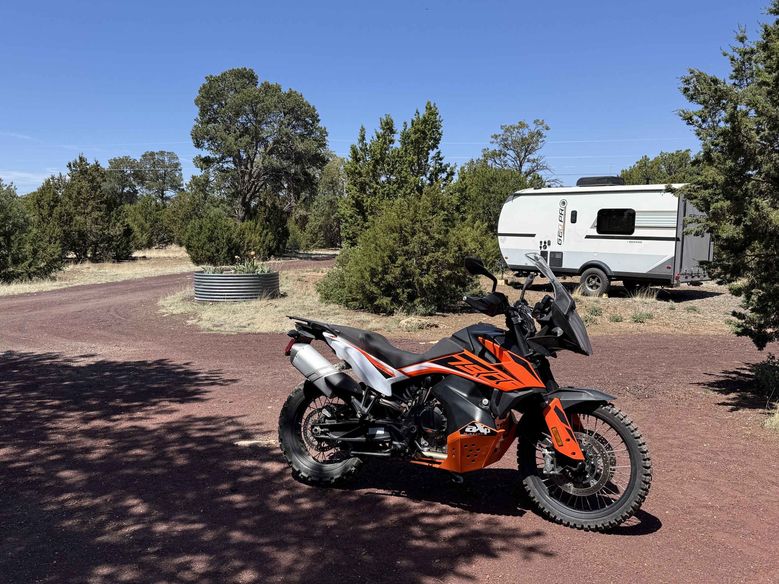 A KTM 790 Adventure motorcycle and a toy hauler trailer in a yard with juniper and pinon pine trees