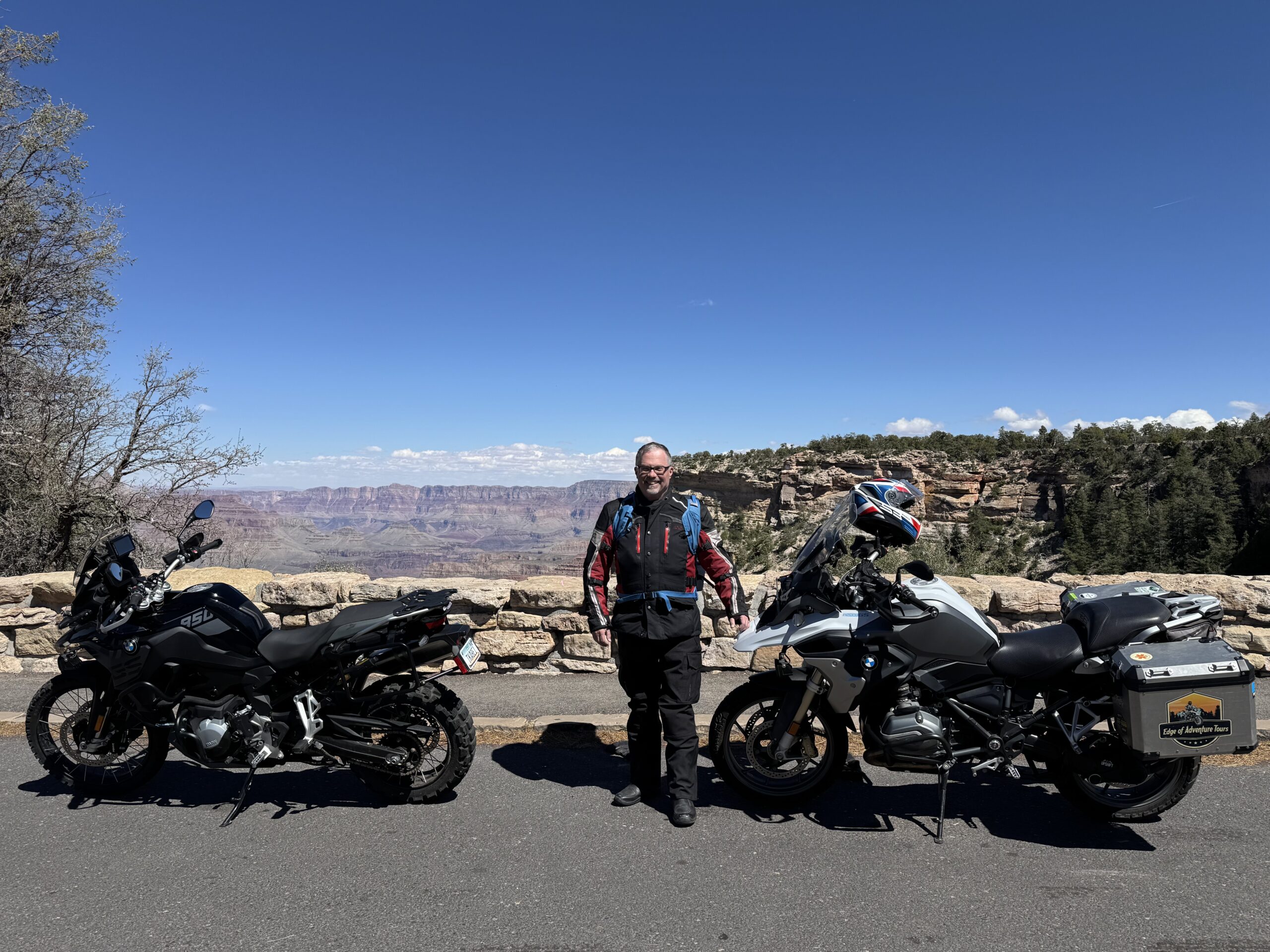 A man stands between a 2023 BMW F850GS and a 2018 BMW R1200GS motorcycle with the Grand Canyon in the background